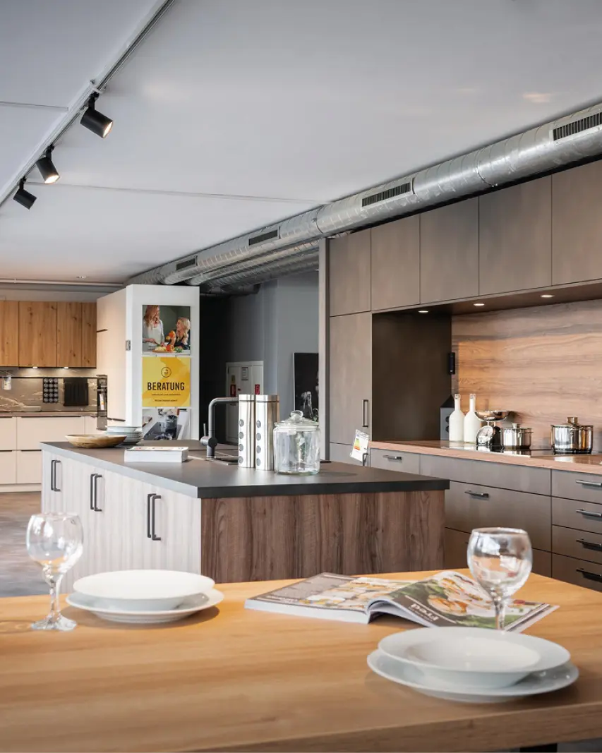 Küchenstudio mit Kochinsel aus Holz und grauer Arbeitsplatte, im Vordergrund Esstisch mit Tellern und Gläsern, im Hintergrund Küchenzeilen